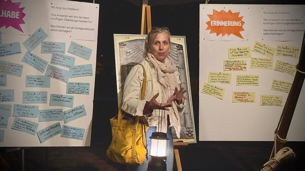 Eine Frau mit einer gelben Tasche steht zwischen zwei Tafeln, die mit bunten Notizen und Texten bedeckt sind, und spricht dabei, wobei sie mit den Händen gestikuliert. Vor ihr steht eine Laterne, die die Szene beleuchtet.