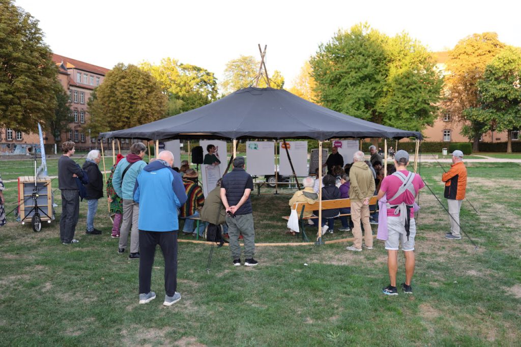 Eine Gruppe von Menschen versammelt sich unter einem großen, runden Vordach in einem Park und betrachtet Informationsplakate, die auf Staffeleien aufgestellt sind.