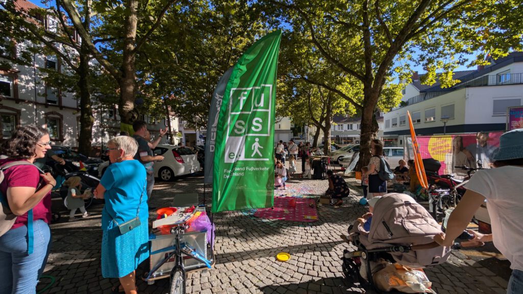 Auf einer sonnendurchfluteten, von Bäumen gesäumten Kopfsteinpflasterstraße versammeln sich Menschen und unterhalten sich in der Nähe eines hohen grünen Banners. Kinderwagen, Fahrräder und bunte Decken sind zu sehen; Gebäude umrahmen diese lebhafte Szene.