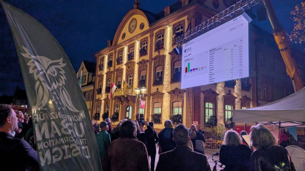 Eine Menschenmenge steht nachts auf einem Platz und beobachtet eine große Leinwand, auf der die Wahlergebnisse an die Fassade eines historischen Gebäudes projiziert werden.