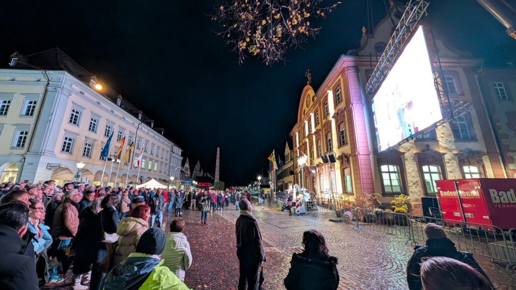 Nachts versammelt sich eine Menschenmenge auf einem Stadtplatz, um auf eine große Videoleinwand im Freien zu schauen.